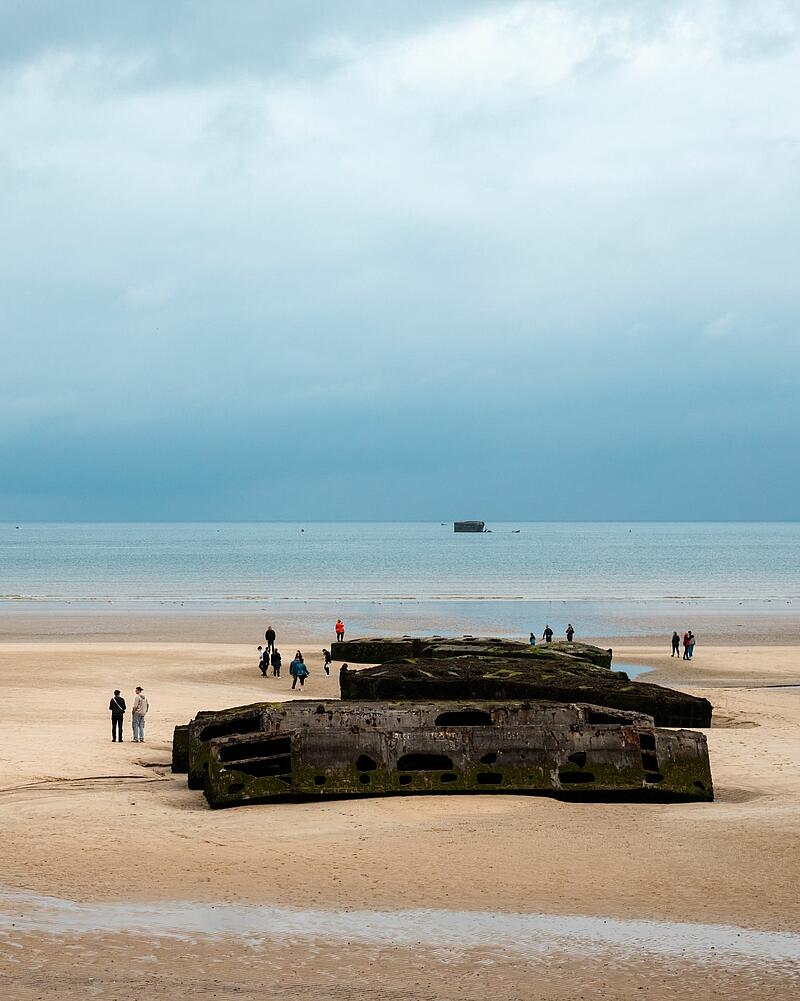 [Translate to Allemand:] Wide shot of a sandy beach under an overcast sky, where tourists walk around large concrete blocks, an excursion easily accessible from Les Castels campings in Normandy.