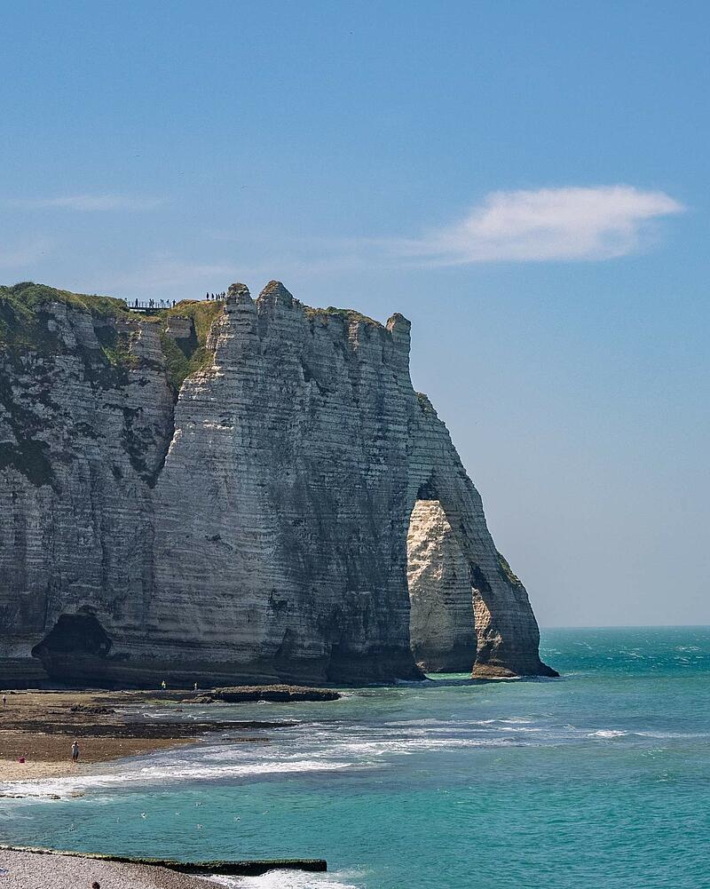 [Translate to Allemand:] A vertical shot of the famous towering white chalk cliffs and natural arch of Étretat, with waves crashing onto the beach, located near a Les Castels camping resort.