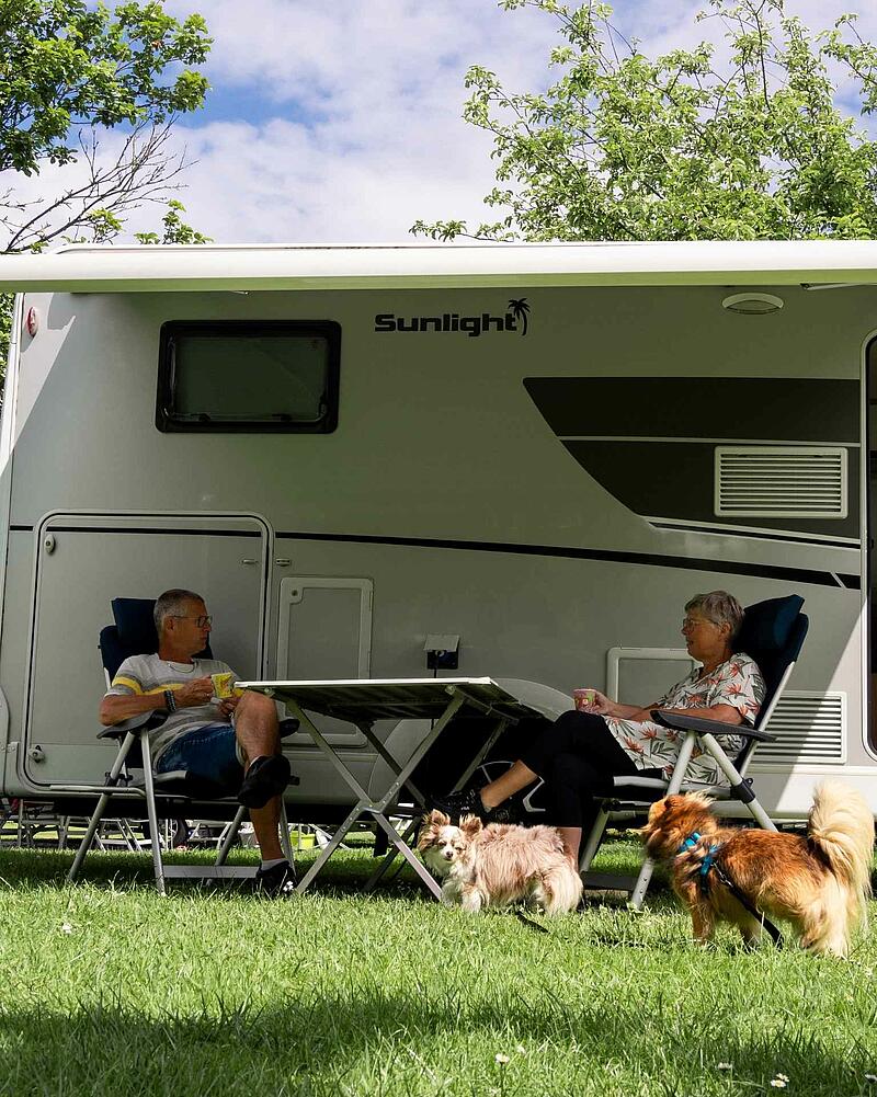 [Translate to Allemand:] A man and a woman sitting at a camping table under the awning of a modern Sunlight motorhome on a pet-friendly pitch at Les Castels.