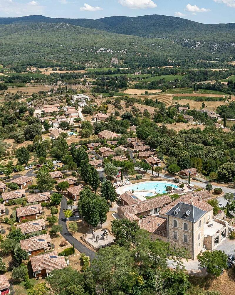 [Translate to Allemand:] Drone perspective of the heated outdoor swimming pool, stone estate building, and eco-friendly wooden chalets surrounded by nature at a 5-star Les Castels campsite.