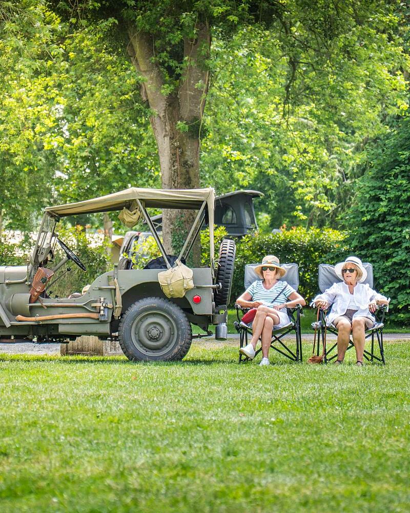 [Translate to Allemand:] Two senior women wearing sun hats sitting on camping chairs on a green lawn next to a classic green vintage military jeep at a Les Castels campsite.