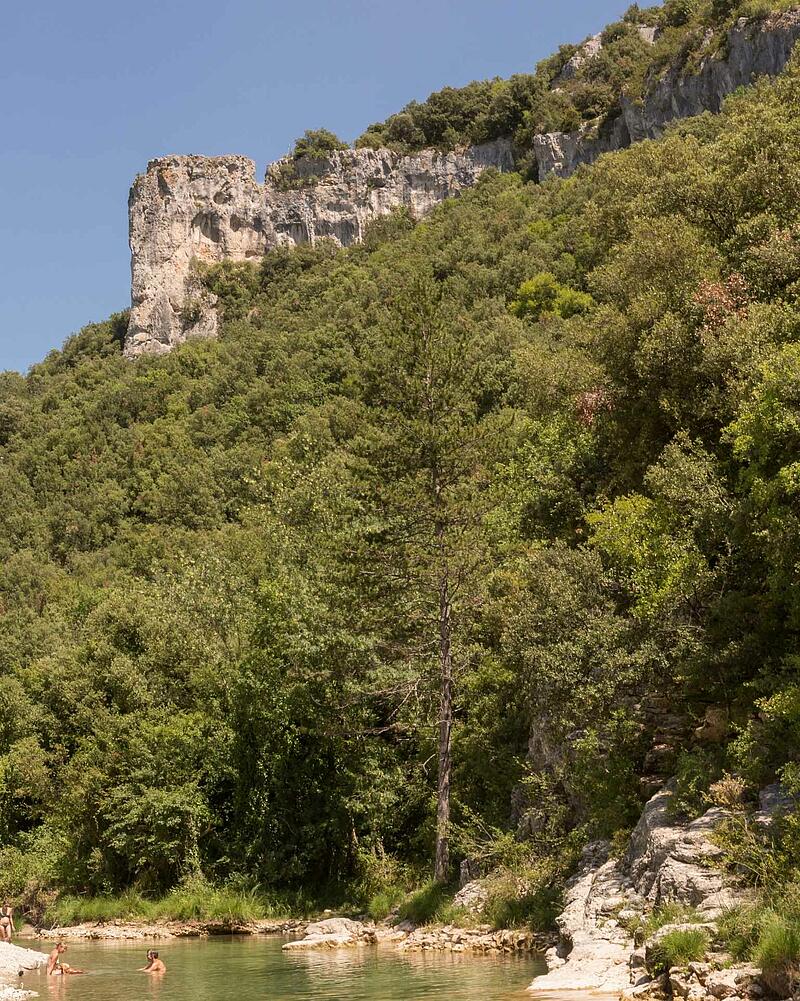 [Translate to Allemand:] People swimming and relaxing in the clear river water at the foot of towering limestone cliffs in the Ardeche Gorges near a luxury Les Castels estate.