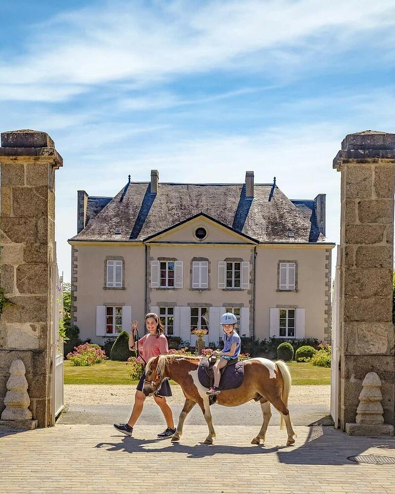 [Translate to Allemand:] A child wearing a riding helmet sits atop a pony led by an instructor, walking past grand stone pillars in front of a majestic historic chateau at a Les Castels campsite.