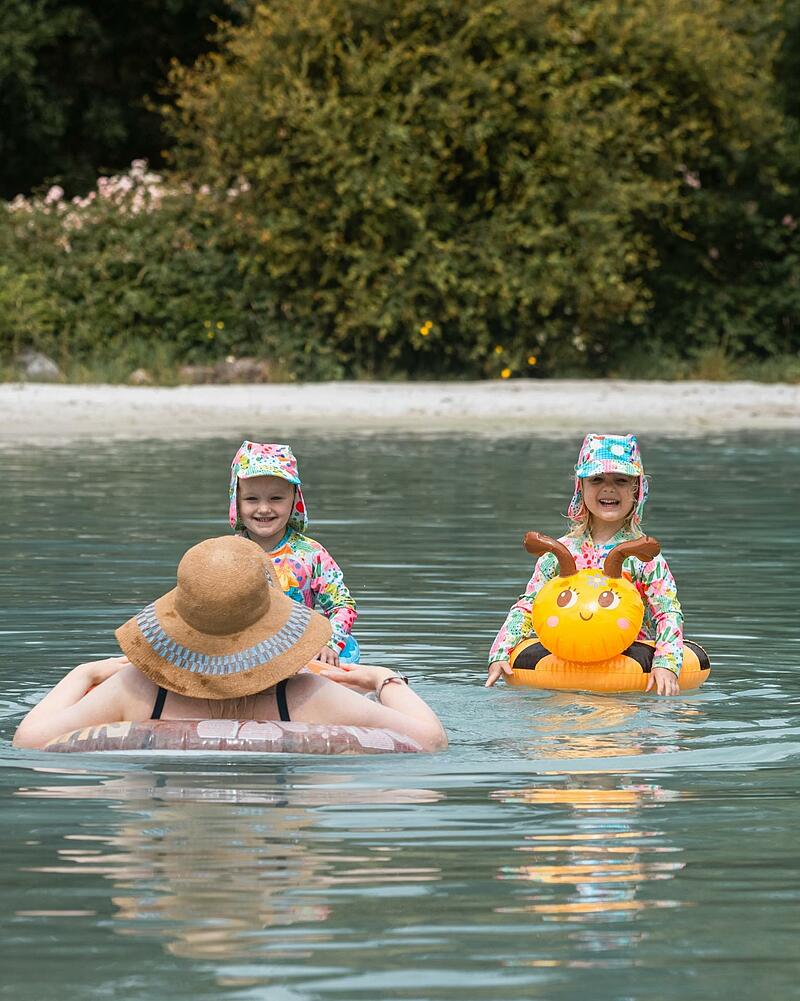 [Translate to Allemand:] Two smiling young children wearing sun-protection swimwear float in the clear turquoise water of a premium Les Castels natural lagoon, alongside an adult wearing a straw hat.