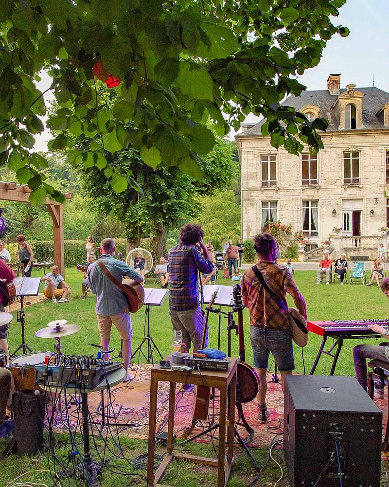 [Translate to Allemand:] Stage view from behind a live band playing guitars and keyboards outdoors for an audience seated on the grass during a premium evening event at Les Castels.