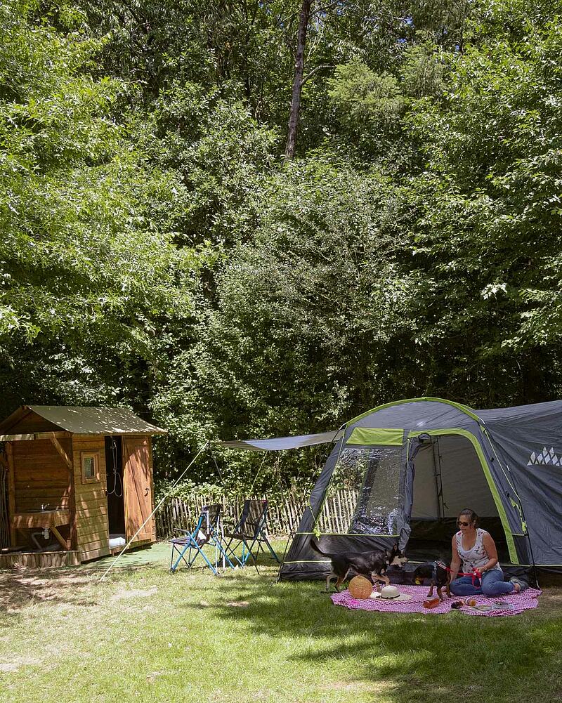 [Translate to Allemand:] A spacious, grassy campsite pitch at a Les Castels campsite, equipped with an exclusive private wooden cabin, and happy pets enjoying the outdoors.