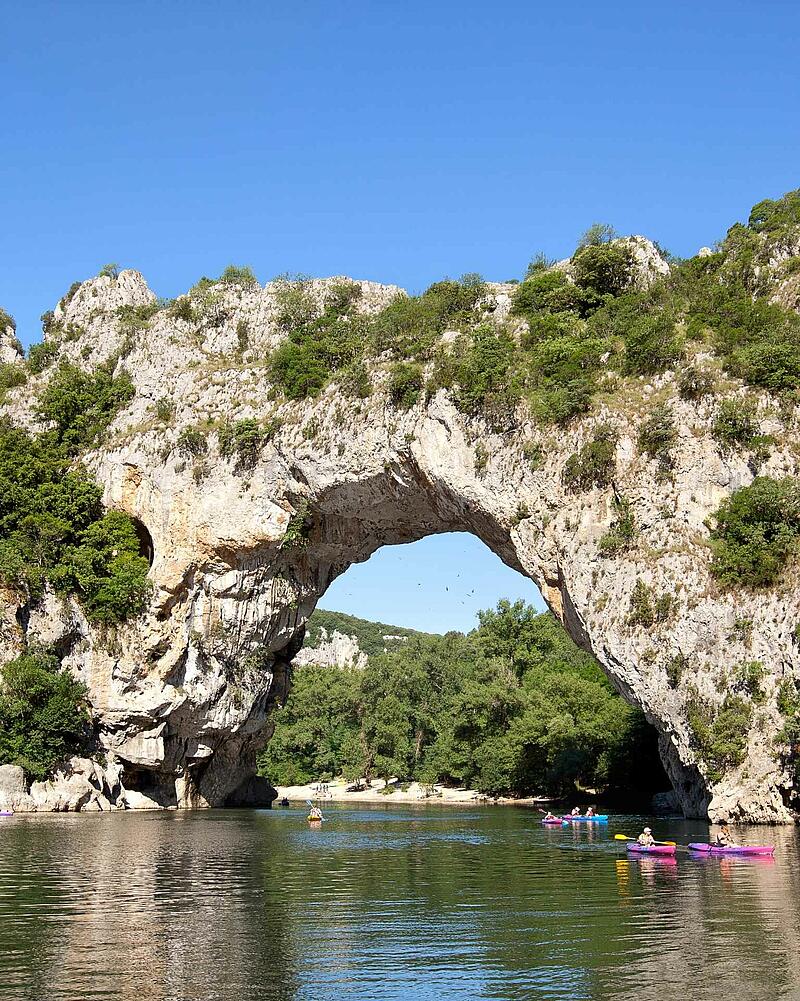 [Translate to Allemand:] Colorful holidaymakers paddling on kayaks and canoes on the river right underneath the giant natural stone bridge of Pont d Arc near Les Castels luxury campsites.