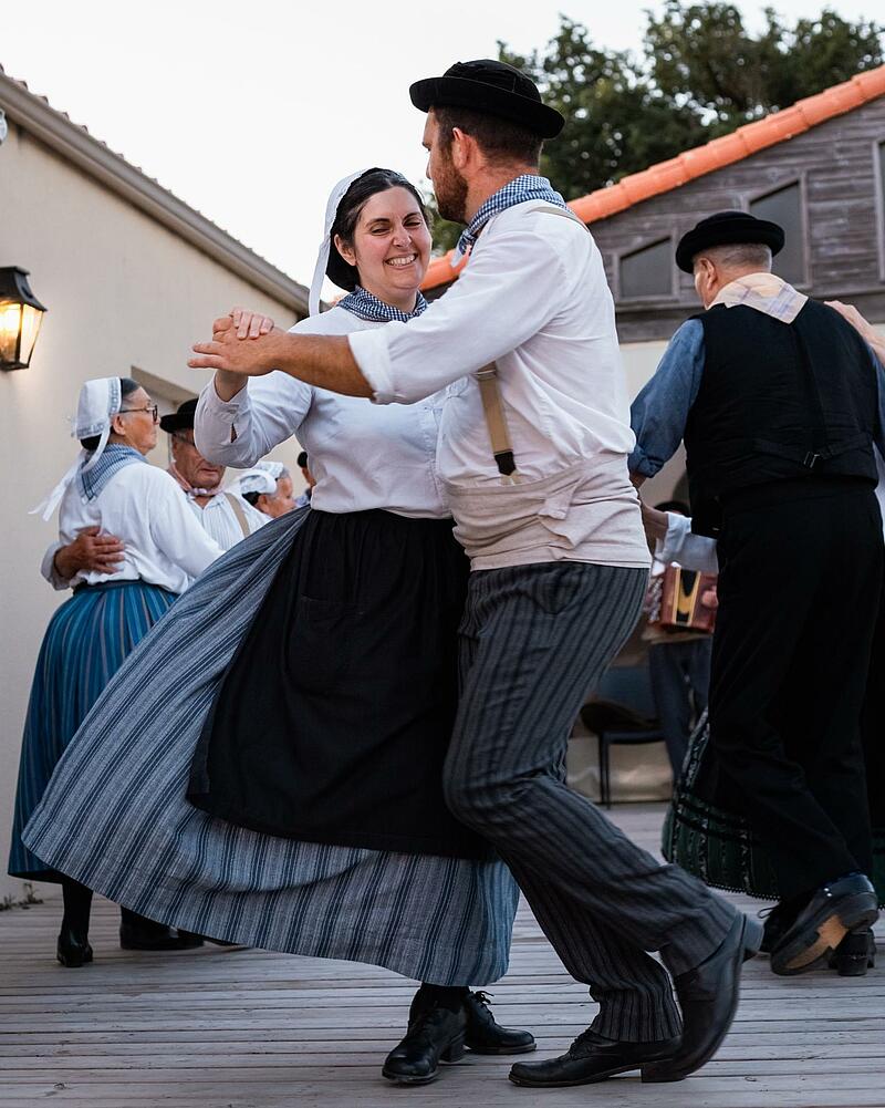 [Translate to Allemand:] Dancers dressed in historic regional attire performing traditional choreography on an outdoor wooden floor during a cultural event at a Les Castels campsite.