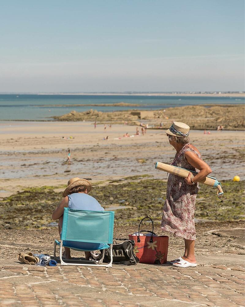 [Translate to Allemand:] Two women wearing straw hats spending a sunny day on a stone seawall, overlooking a vast sandy beach during a seaside holiday with Les Castels campings.