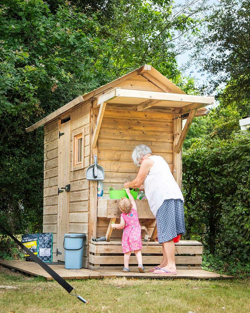 [Translate to Allemand:] Woman and young girl interact at a rustic wooden outdoor kitchen station on a Les Castels camping estate.