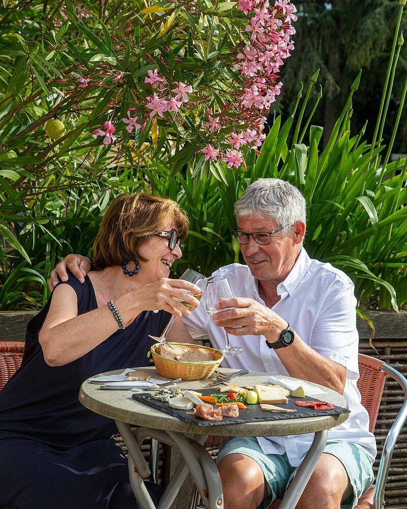 [Translate to Allemand:] A man and a woman laughing and clinking wine glasses at an outdoor table, enjoying an artisanal cheese board at a premium Les Castels campsite restaurant.