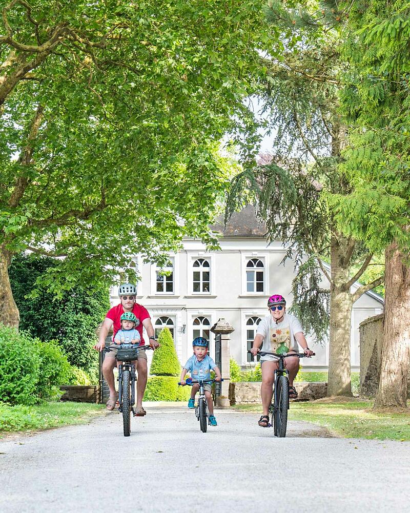 [Translate to Allemand:] A family of four, including two young children wearing helmets, cycling towards the camera on a gravel path during their holiday at a Les Castels domain.