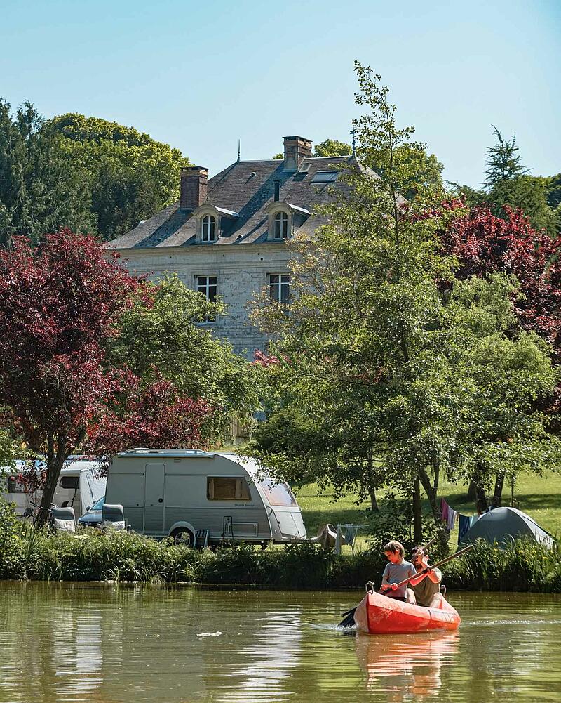 [Translate to Allemand:] An orange canoe with a man and a young boy paddling on a calm lake, with a caravan parked on the grassy bank of a Les Castels estate.