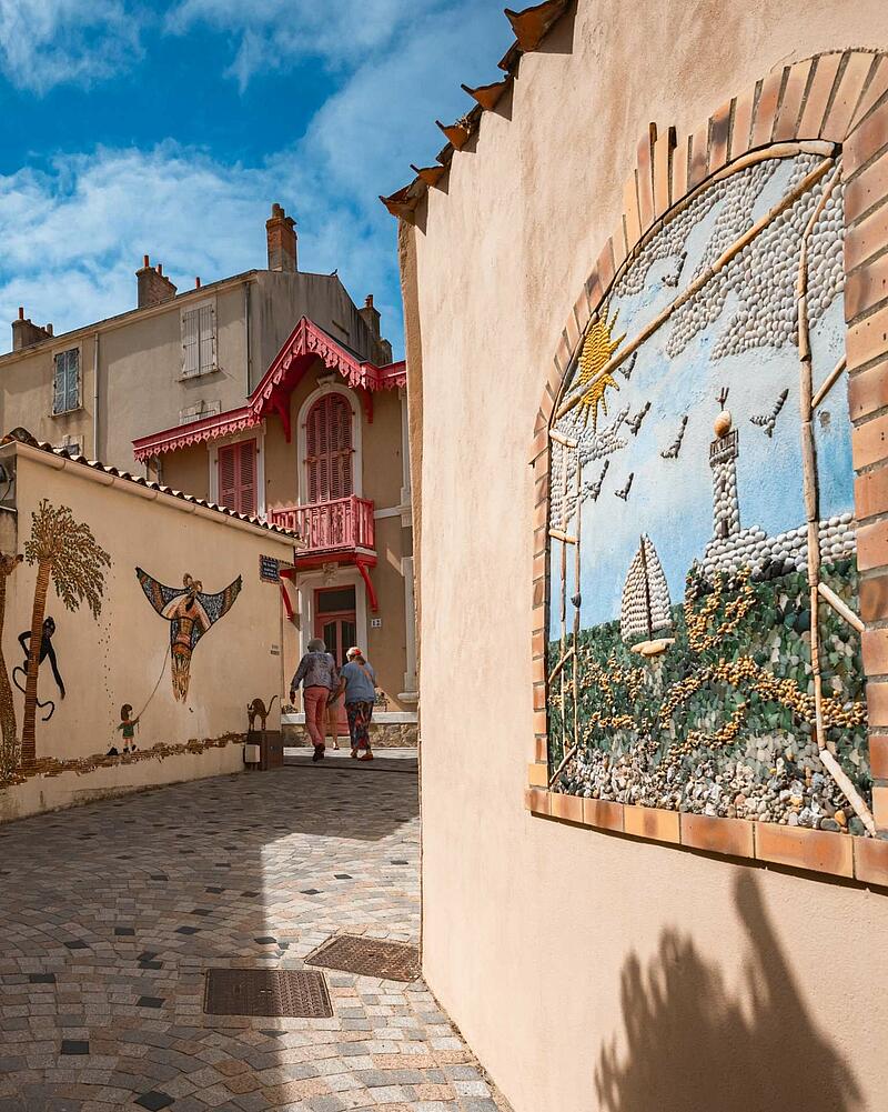 [Translate to Allemand:] A charming European street in Île Penotte, close to a Les Castels campsite, featuring vibrant shell mosaic artwork on the walls of light-colored buildings.