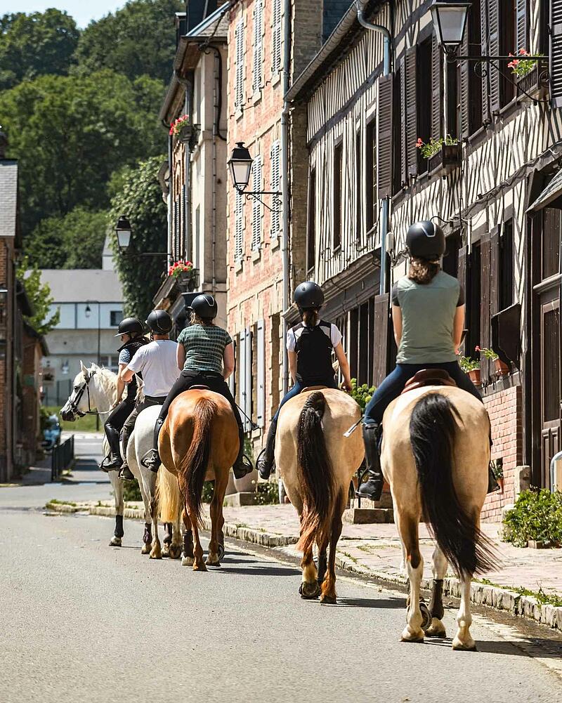 [Translate to Allemand:] A group of riders on horseback walking down a village street lined with classic half-timbered stone houses, an outdoor activity proposed near your Les Castels camping resort.