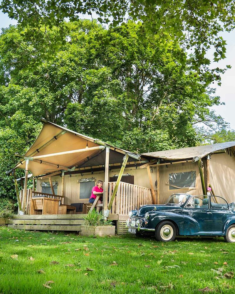 [Translate to Allemand:] A large canvas glamping safari tent with a wooden porch set amongst lush green trees on a premium pitch at a Les Castels 5-star campsite.