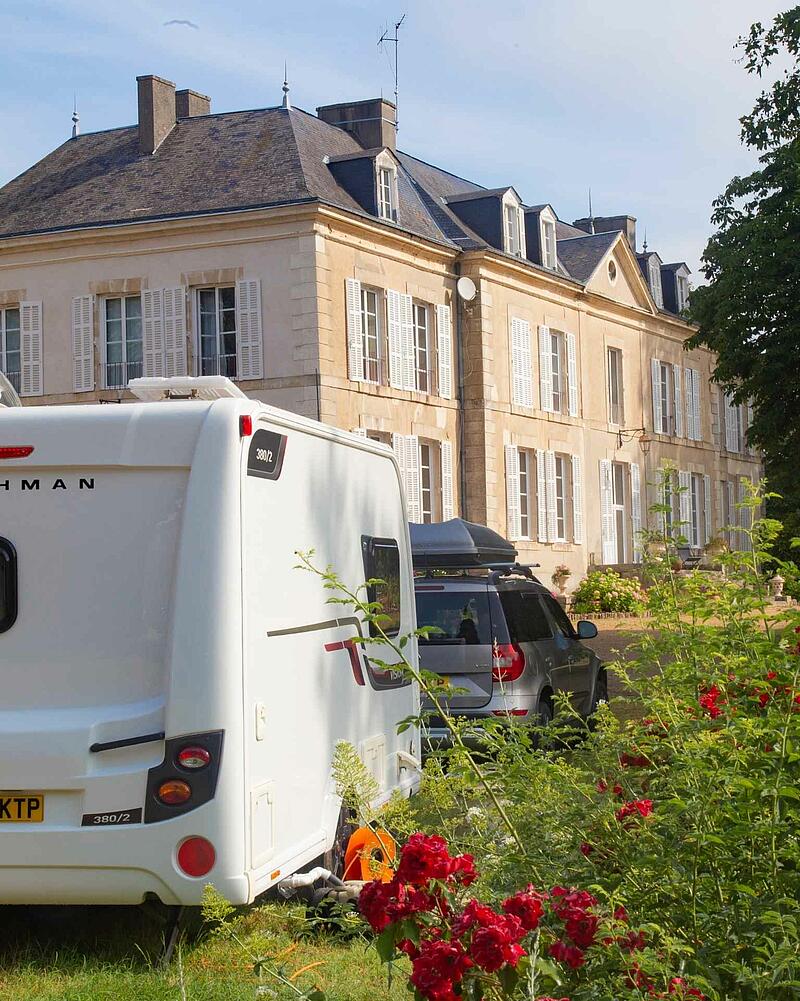 [Translate to Allemand:] The back of a white caravan and a grey SUV parked on a sunny grass pitch in front of a grand, historic stone chateau, beautifully framed by blooming red roses in the foreground.