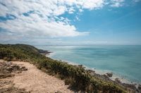[Translate to Allemand:] A scenic coastal view in Normandy featuring a dirt pathway on a grassy cliff overlooking a vast expanse of turquoise sea under a blue sky with fluffy white clouds, perfect for an outdoor stay at Les Castels.